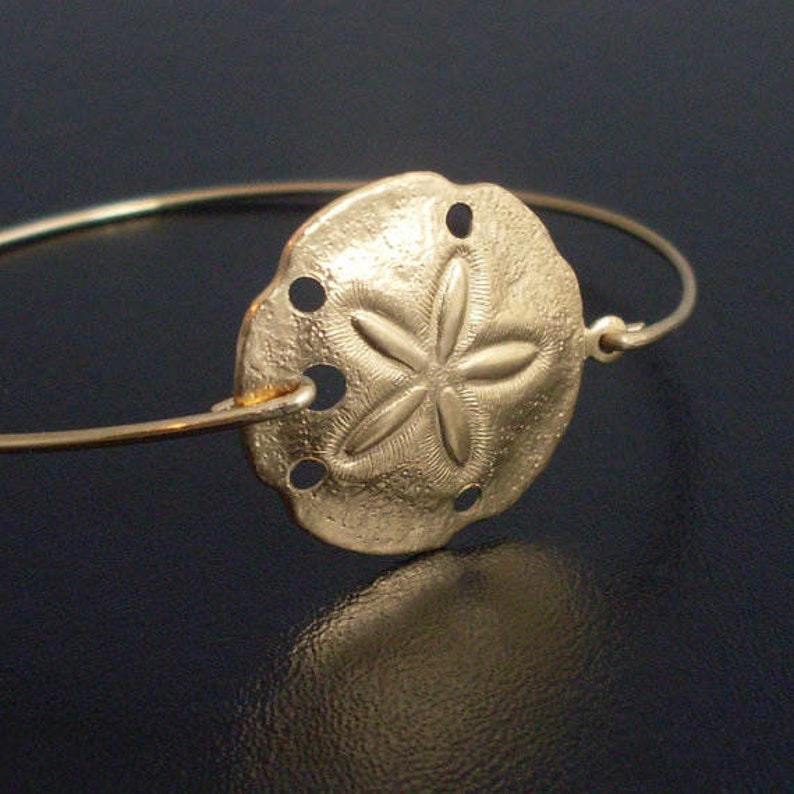 May include: A gold-toned bangle bracelet with a sand dollar charm. The charm is detailed with a five-pointed star and textured surface.