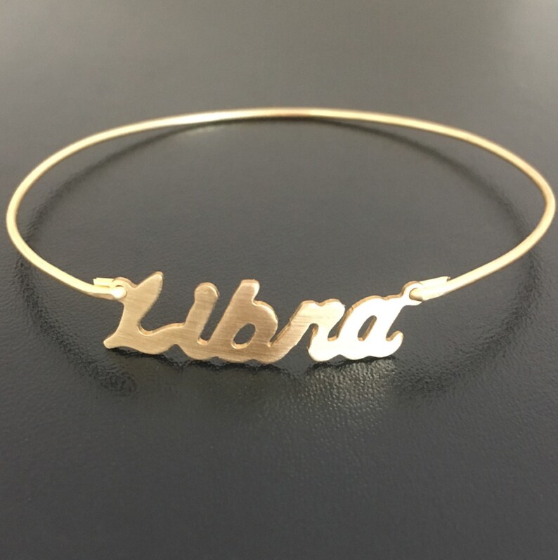 May include: Gold bangle bracelet with the word "Libra" written in a cursive font.