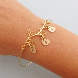 May include: Gold bracelet with a tree branch design and three heart charms. The charms are engraved with the letters "S", "K", and "E".