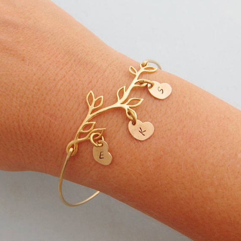 May include: Gold bracelet with a tree branch design and three heart charms. The charms are engraved with the letters "S", "K", and "E".