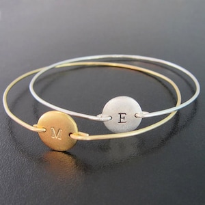 May include: Two gold and silver bangle bracelets with engraved initial charms. The gold bracelet has an "M" charm and the silver bracelet has an "E" charm.