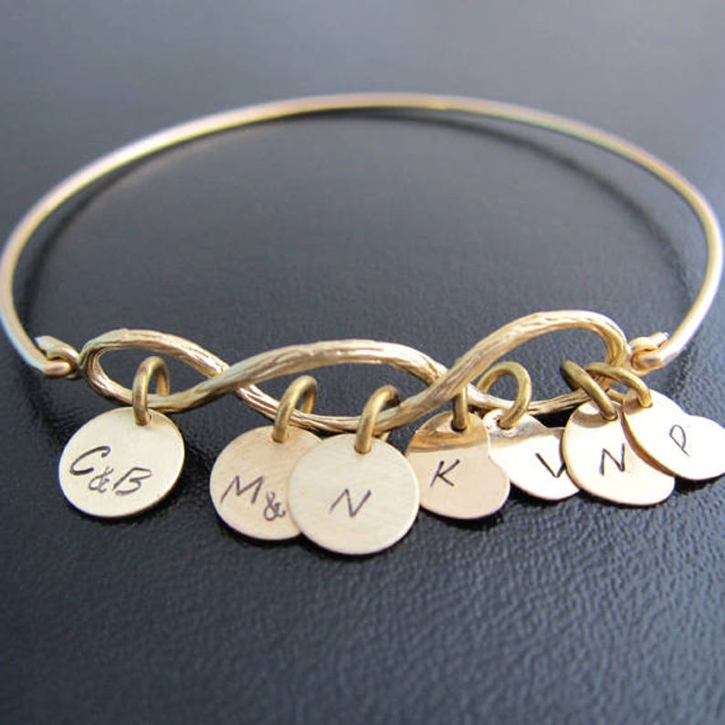 May include: Gold infinity bangle bracelet with multiple charms. Each charm is a small round disc with initials engraved on them. The charms include the initials C&B, M&N, N, K, V, N, and P.