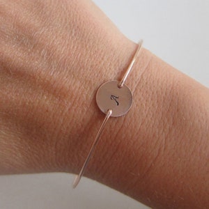 May include: A rose gold bangle bracelet with a round charm engraved with the letter "A".