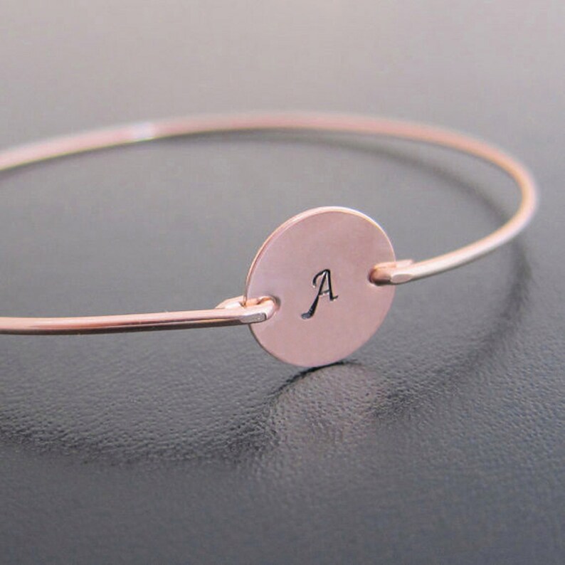 May include: A rose gold bangle bracelet with a round charm featuring the letter 'A' engraved in a cursive font.
