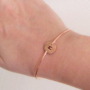 K&ouml;nnte beinhalten: Ein zartes ros&eacute;goldenes Armband mit einem d&uuml;nnen Band und einem kreisf&ouml;rmigen Anh&auml;nger. Der Anh&auml;nger ist mit dem Buchstaben "M" in einer schlichten, eleganten Schrift graviert. Das Armband ist f&uuml;r das Handgelenk konzipiert.