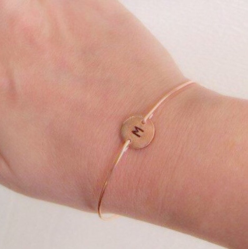K&ouml;nnte beinhalten: Ein zartes ros&eacute;goldenes Armband mit einem d&uuml;nnen Band und einem kreisf&ouml;rmigen Anh&auml;nger. Der Anh&auml;nger ist mit dem Buchstaben "M" in einer schlichten, eleganten Schrift graviert. Das Armband ist f&uuml;r das Handgelenk konzipiert.
