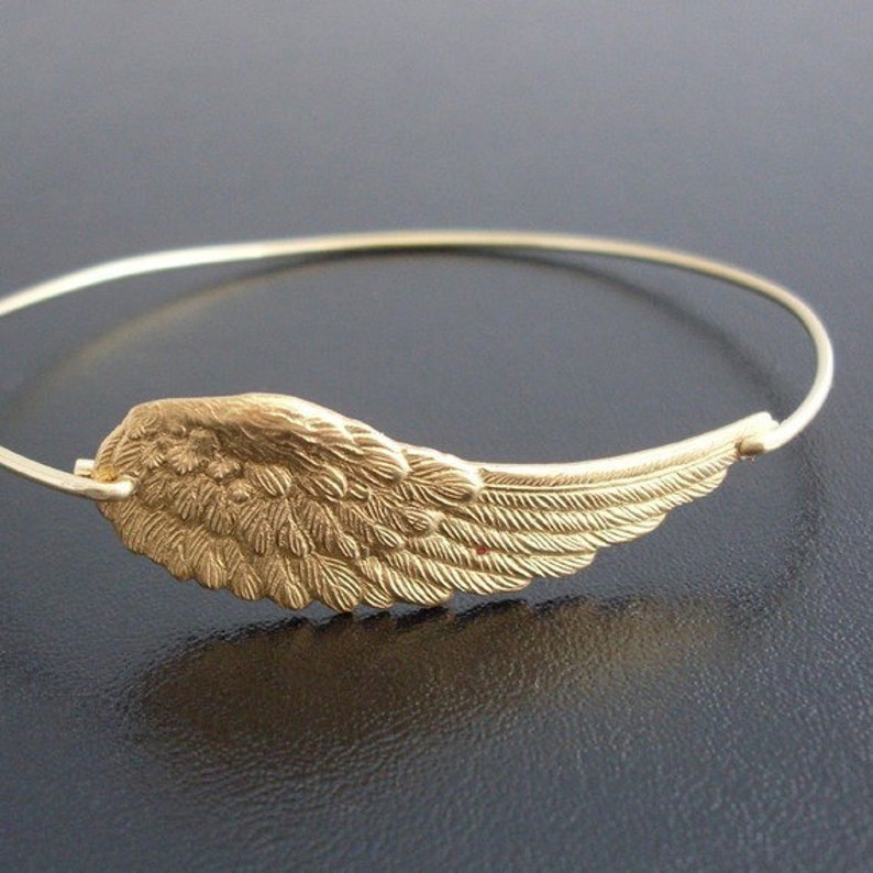 May include: A gold-toned bangle bracelet with a detailed angel wing charm. The wing is textured and has a delicate, feminine design.