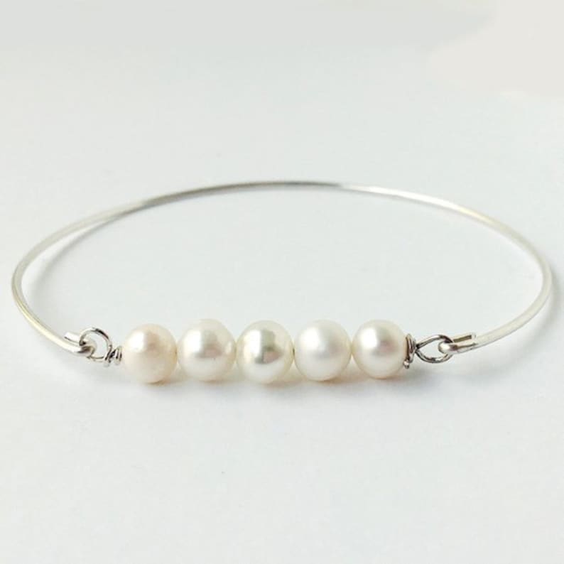 K&ouml;nnte beinhalten: Ein filigranes silbernes Armreif-Armband mit f&uuml;nf gl&auml;nzenden, cremefarbenen Perlen. Die Perlen sind gleichm&auml;&szlig;ig auf der Vorderseite des Armbands angeordnet und schaffen ein schlichtes, aber elegantes Design. Das Armband hat einen klassischen, minimalistischen Stil.