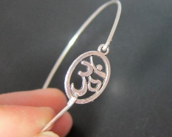 May include: A silver-colored bangle bracelet featuring an oval charm with an Om symbol. The bracelet has a simple, minimalist design, with a thin, curved band. The charm is intricately detailed, showcasing the Om symbol's unique shape.