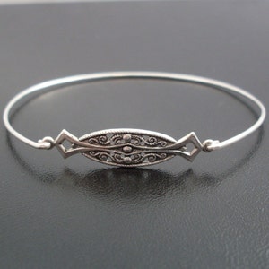 May include: A silver bangle bracelet with a decorative filigree design. The design features a central oval shape with intricate swirls and a beaded border.