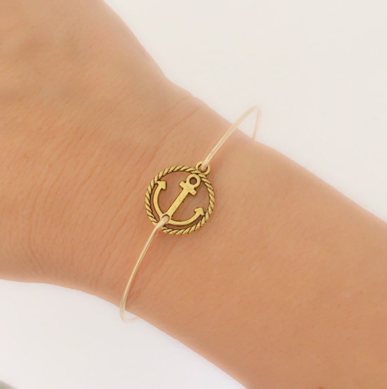 K&ouml;nnte beinhalten: Ein goldfarbenes Anker-Armband mit einem d&uuml;nnen, durchsichtigen Drahtband. Der Anker ist von einem Seil-Design umgeben.