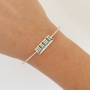 May include: A silver bangle bracelet with a piano key design charm. The bracelet is adjustable and has a simple, elegant design.