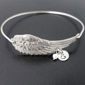 May include: A silver bangle bracelet with a detailed angel wing charm and a small pearl and initial charm. The initial charm is engraved with the letter "E".