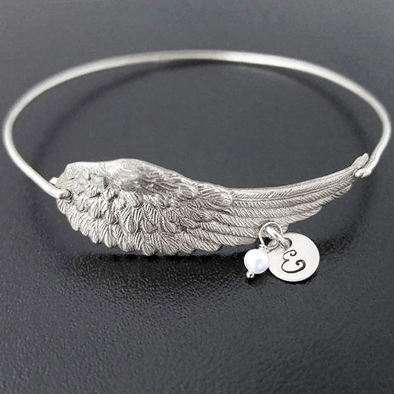 May include: A silver bangle bracelet with a detailed angel wing charm and a small pearl and initial charm. The initial charm is engraved with the letter "E".