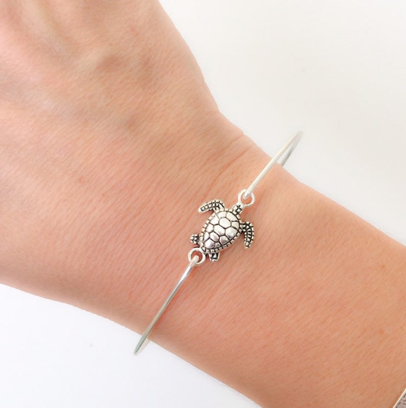 May include: A silver bangle bracelet with a silver turtle charm. The bracelet is on a person's wrist.