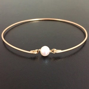 May include: A gold-colored bangle bracelet featuring a single, round, white pearl at the center. The bracelet has a simple, elegant design with a clasp closure. The pearl is approximately 0.3 inches in diameter.