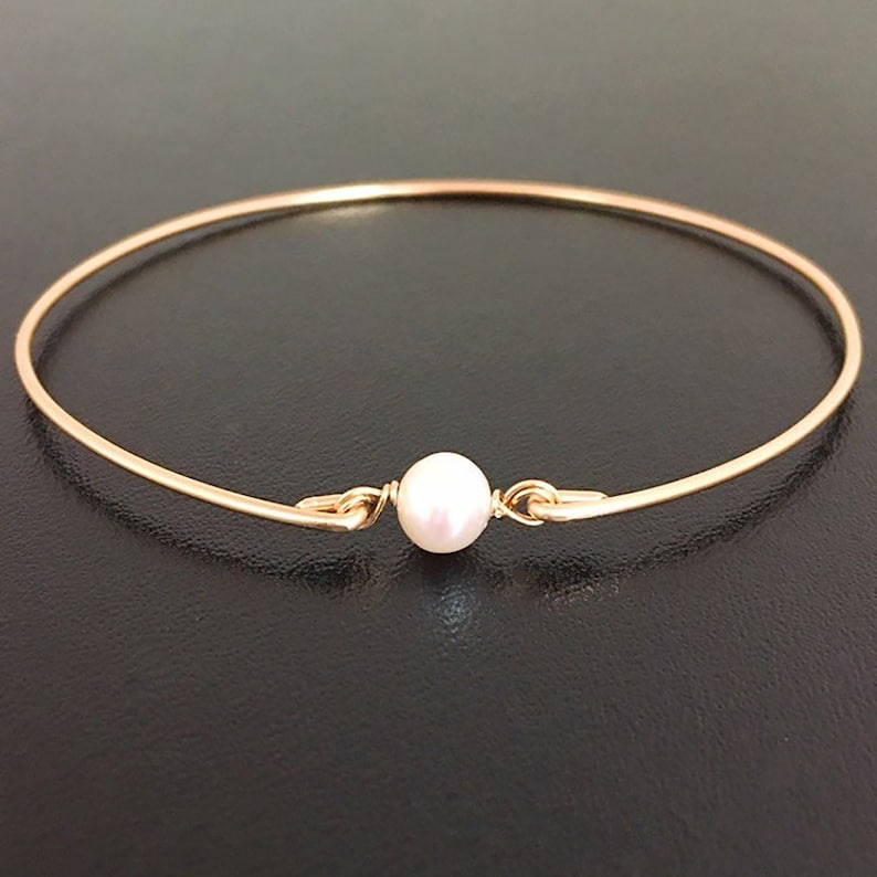 May include: A gold-colored bangle bracelet featuring a single, round, white pearl at the center. The bracelet has a simple, elegant design with a clasp closure. The pearl is approximately 0.3 inches in diameter.