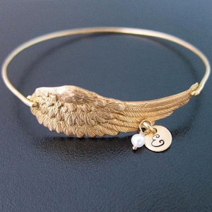 May include: A gold bangle bracelet with a detailed angel wing charm and a small pearl bead. The charm has a small disc with the letter "E" engraved on it.