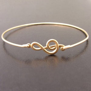 May include: A gold-toned bangle bracelet with a treble clef charm. The bracelet is thin and delicate.