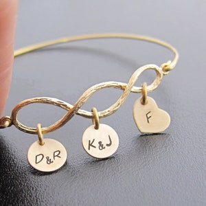 May include: Gold infinity bracelet with three charms. The charms are round and engraved with the initials "D&R", "K&J", and a heart charm with the letter "F".