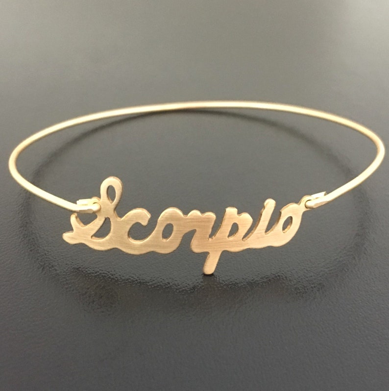 May include: Gold bangle bracelet with the word "Scorpio" written in a cursive font.