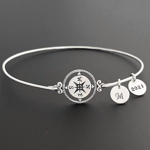 May include: A silver bangle bracelet with a compass charm and two additional charms. One charm is engraved with the letter "M" and the other with the year "2021".
