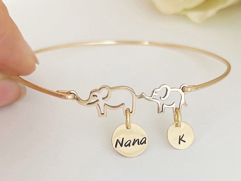 May include: Gold bangle bracelet with two elephant charms and two round charms. The larger charm says "Nana" and the smaller charm says "K".