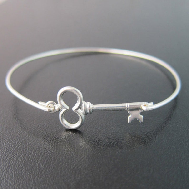 Peut inclure: Un bracelet argent&eacute; avec un charme en forme de cl&eacute;. Le charme est une cl&eacute; stylis&eacute;e avec un n&oelig;ud en forme de c&oelig;ur.