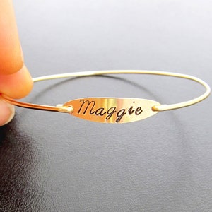 May include: A gold bangle bracelet with a small oval charm engraved with the name "Maggie".