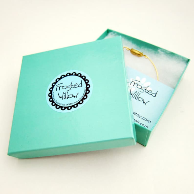 Puede incluir: Una caja de regalo turquesa con una etiqueta blanca que dice "Frosted Willow" y una direcci&oacute;n de sitio web. La caja est&aacute; abierta y un brazalete de alambre dorado est&aacute; dentro.