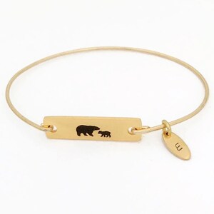 May include: Gold bangle bracelet with a rectangular charm featuring a silhouette of a mother bear and cub. The charm has a small tag with the letter "E" engraved on it.