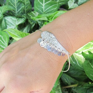 May include: Silver bracelet with a wing charm and multiple charms with letters on them. The bracelet is on a person's wrist.
