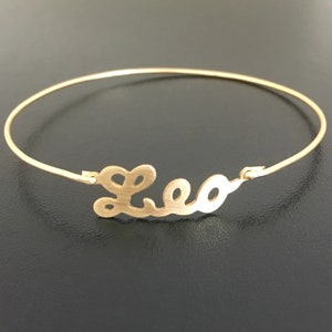 May include: A gold-toned bangle bracelet with the name "Leo" cut out in a cursive font.