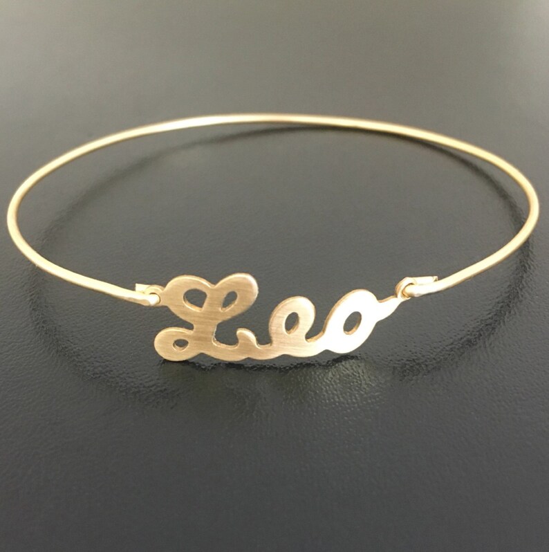 May include: A gold-toned bangle bracelet with the name "Leo" cut out in a cursive font.