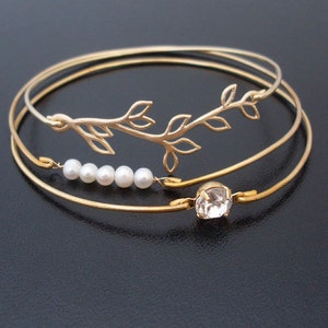 May include: A set of three gold bangle bracelets with a delicate branch design and a single clear rhinestone charm. The bracelets are adorned with five small white pearls.