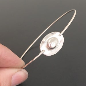 May include: A silver bangle bracelet with a round, textured silver charm. The charm has a raised, smooth, oval-shaped center.
