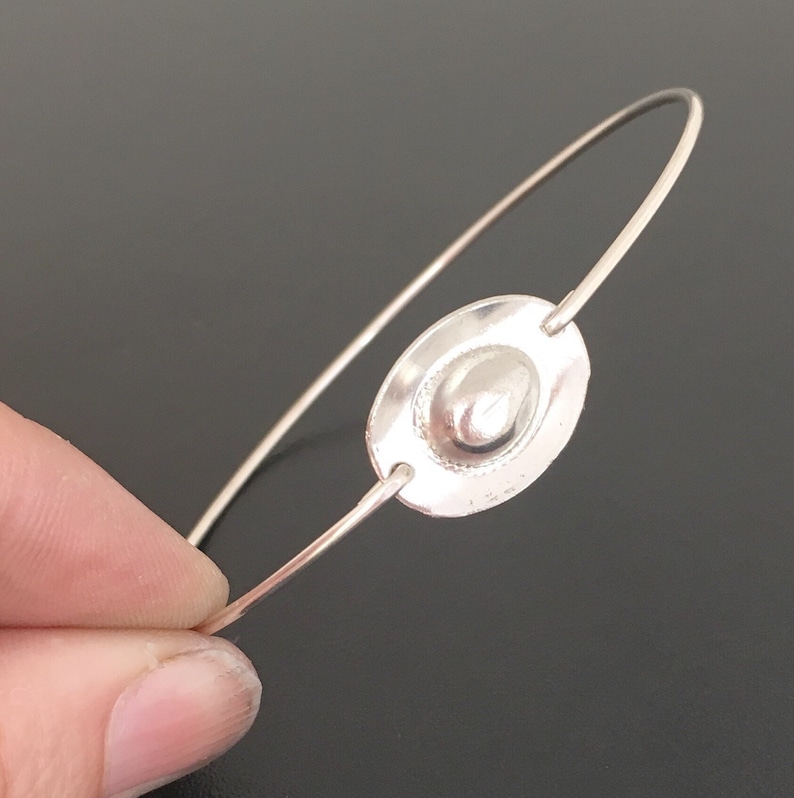 May include: A silver bangle bracelet with a round, textured silver charm. The charm has a raised, smooth, oval-shaped center.