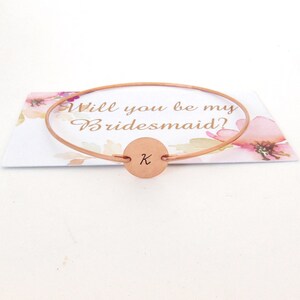 Puede incluir: Pulsera de dama de honor de oro rosa con un dije redondo grabado con la letra K. La pulsera est&aacute; sobre una tarjeta que dice "&iquest;Quieres ser mi dama de honor?"
