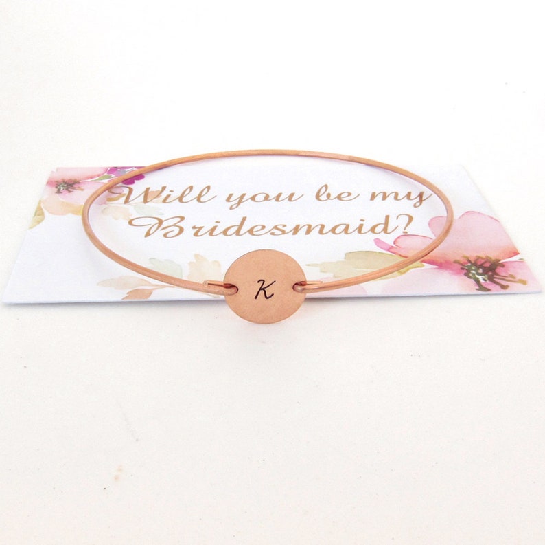 Puede incluir: Pulsera de dama de honor de oro rosa con un dije redondo grabado con la letra K. La pulsera est&aacute; sobre una tarjeta que dice "&iquest;Quieres ser mi dama de honor?"