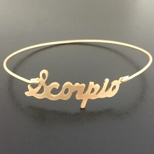 Pode incluir: Pulseira de bracelete de ouro com a palavra "Scorpio" escrita em cursiva.