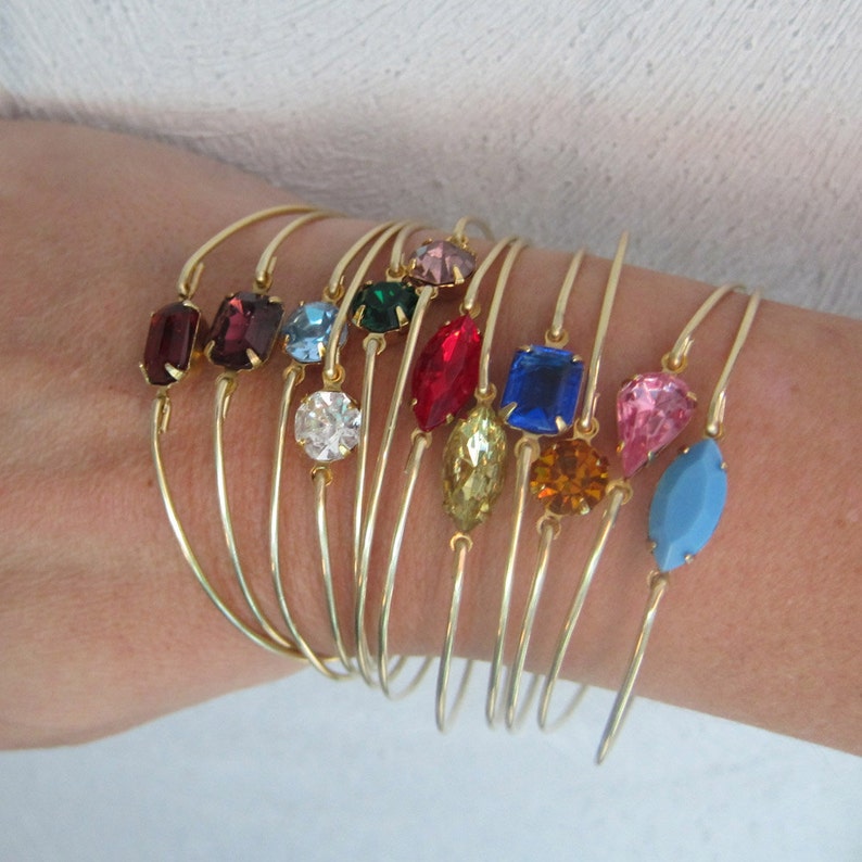 May include: A set of gold bangle bracelets with colorful gemstone accents. The bracelets feature a variety of shapes and sizes of gemstones, including round, square, and oval. The gemstones are set in gold settings and are surrounded by a thin gold band.