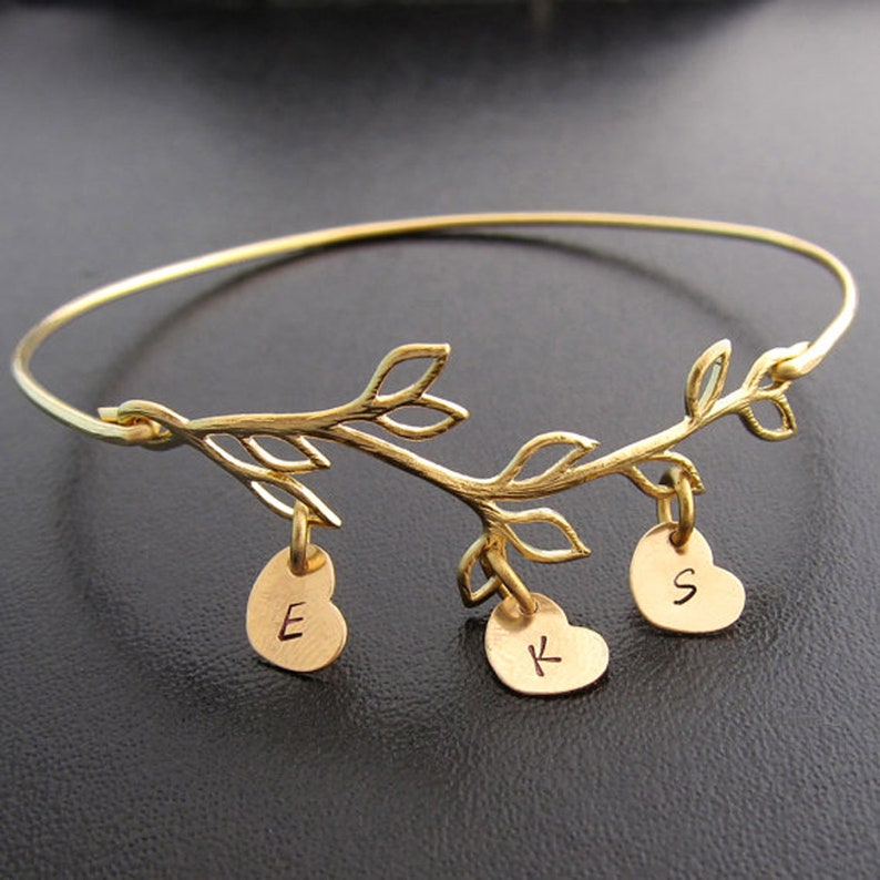 May include: Gold bangle bracelet with a delicate branch design and three heart-shaped charms. The charms are engraved with the letters "E", "K", and "S".