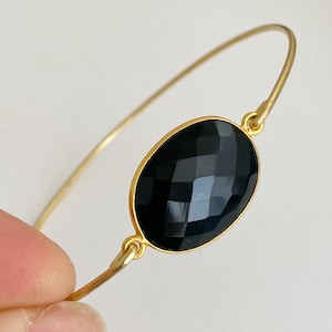 May include: A gold bangle bracelet with a large, oval-shaped black onyx gemstone. The gemstone has a faceted cut.