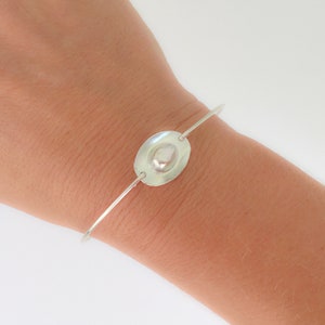 May include: A silver bangle bracelet with a smooth, oval-shaped charm. The charm has a raised, textured center.