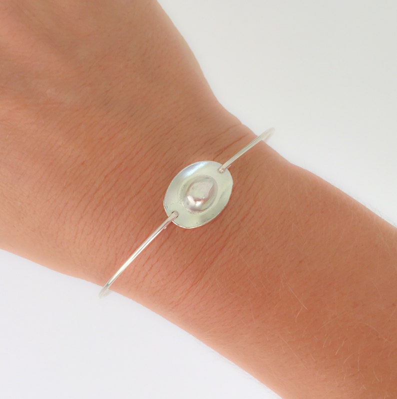 May include: A silver bangle bracelet with a smooth, oval-shaped charm. The charm has a raised, textured center.
