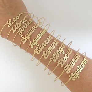 May include: A set of twelve gold-toned bangle bracelets, each featuring a different zodiac sign in a cursive script. The bracelets are stacked on a person's arm.
