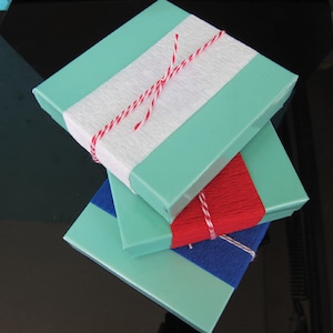 May include: Three square boxes stacked on top of each other. The top box is turquoise with a white stripe and is tied with red and white twine. The middle box is turquoise and the bottom box is blue.