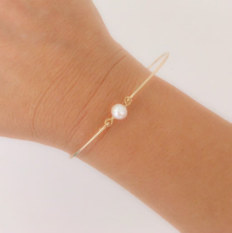 May include: A delicate gold bangle bracelet with a single white pearl accent. The bracelet is simple and elegant, perfect for everyday wear.