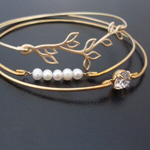 Pode incluir: Tr&ecirc;s pulseiras de ouro com um delicado design de folhas, uma fileira de p&eacute;rolas e um strass transparente.