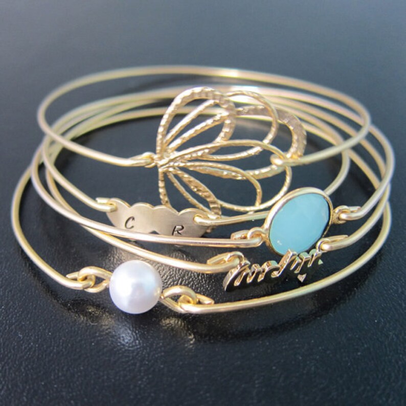 May include: A collection of gold-colored bangles. One bangle features a butterfly design, another has a cloud-shaped charm with the letters "C R", and another has a light blue oval stone. A fourth bangle has a pearl, and the last one has the word "forever".